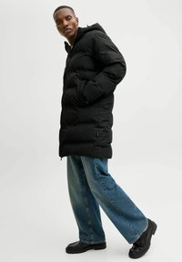 Schwarze Pufferjacke mit Kapuze, Reißverschluss und Seitentaschen; kombiniert mit locker sitzenden, hellblauen Jeans und schwarzen Stiefeln.