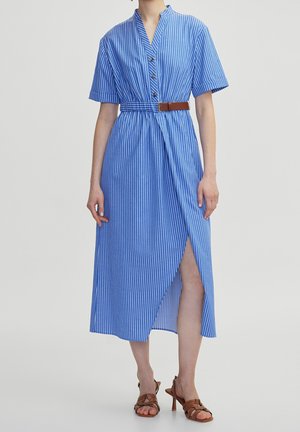 Femme portant une robe midi rayée verticale bleue et blanche avec manches courtes, ceinture marron, boutons sur le devant, fente latérale et sandales à talons marron.