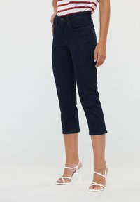 Jean en denim foncé, coupé, avec une silhouette ajustée, des poches avant et une fermeture à bouton. Associé à des sandales à talons hauts blanches avec des lanières.