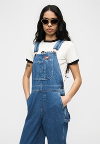 Γυναίκα με μακριά σκούρα μαλλιά που φοράει γυαλιά ηλίου, άσπρο T-shirt και μπλε τζιν φορμάκι, με τα χέρια στις τσέπες, στέκεται μπροστά σε απλό φόντο.