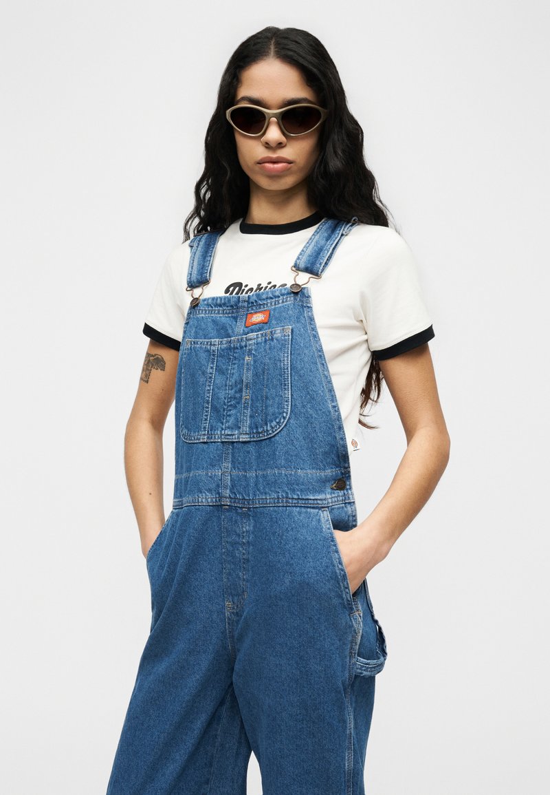 Donna con capelli lunghi e scuri che indossa occhiali da sole, una maglietta bianca e una salopette di jeans blu con le mani nelle tasche, in piedi contro uno sfondo semplice.