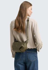 Olivengrüne Leder-Schultertasche mit strukturiertem Finish, verstellbarem Tragegurt und Kontrastnähten. Vor einem neutralen Hintergrund getragen.