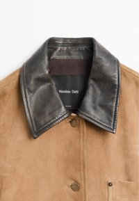 Giacca scamosciata beige con collo in pelle nera e chiusura frontale a scatto, dotata di tasca laterale e etichetta del marchio visibile all'interno.