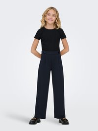 Jeune fille aux cheveux bouclés blonds portant un haut noir à manches courtes, un pantalon large bleu marine avec des détails à boutons, et des mocassins noirs, souriante.