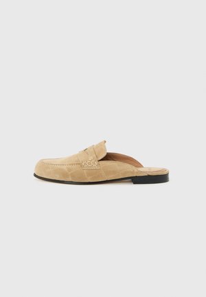 Beige ruskind backless loafer med sort sål og synlige sømme på en ensfarvet lys baggrund.
