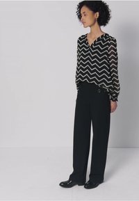 Blusa a motivo a chevron in bianco e nero abbinata a pantaloni neri a gamba larga. Materiale trasparente con maniche lunghe, vita aderente con dettagli laterali.
