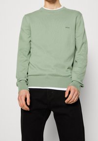 Homme portant un pull vert clair à col rond avec un petit logo "BOSS" sur la poitrine et un pantalon noir, debout devant un fond uni.