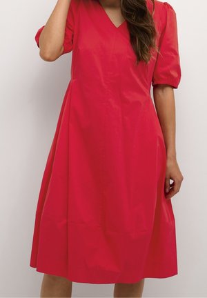 Femme portant une robe rouge longueur genou avec des manches courtes bouffantes et un décolleté en V, debout devant un fond clair uni.