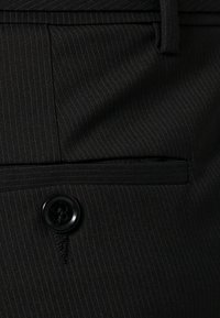 Černá látka s pruhy, která má přední kapsu a černý knoflíkový detail. Materiál má hladkou texturu a formální vzhled.
