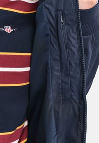 Marinejakke med en lynlås lomme og ribbede manchetter, lagd over en bordeaux, navy og hvid stribet skjorte med et logo på brystet.