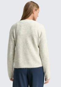 O femeie cu păr lung poartă un pulover gri deschis cu model în relief și o pereche de pantaloni albaștri închiși, întoarsă cu spatele față de un fundal simplu și deschis.