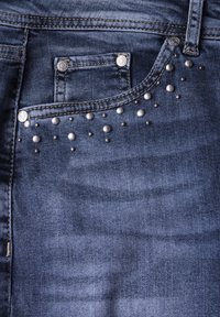 Jean en denim bleu foncé avec une poche avant ornée d'accents en métal argenté disposés en motif près de la poche et de la taille.