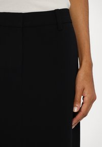 Pantaloni negri, croiți, dintr-un material neted, având un tiv plat și cusături laterale. Mână odihnindu-se ușor pe material.