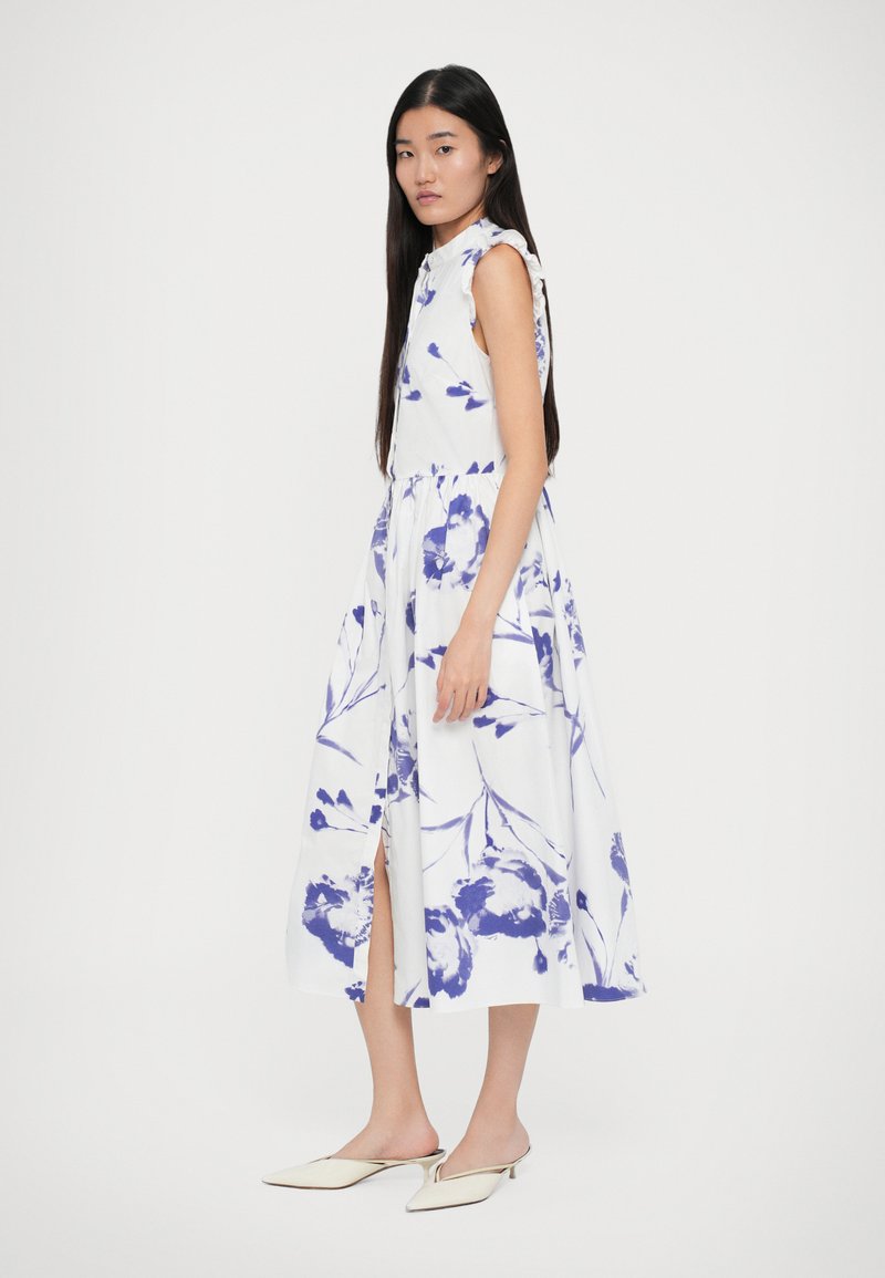 White floral dress featuring purple patterns, sleeveless design, and a gathered waist. Includes a side slit and is paired with cream-colored mules.
