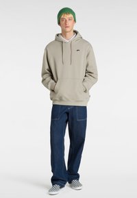 Grå hoodie med framficka, kombinerad med lösa mörka denimjeans och rutig slip-on skor. Modellen bär en grön stickad mössa.