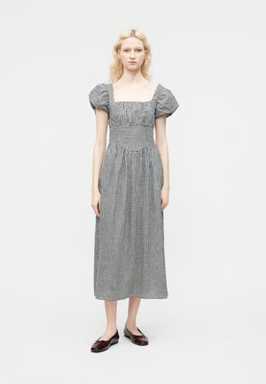 Blonde Frau trägt ein schwarz-weiß kariertes Midikleid mit Puffärmeln und quadratischem Ausschnitt, kombiniert mit dunkelroten Ballerinas.