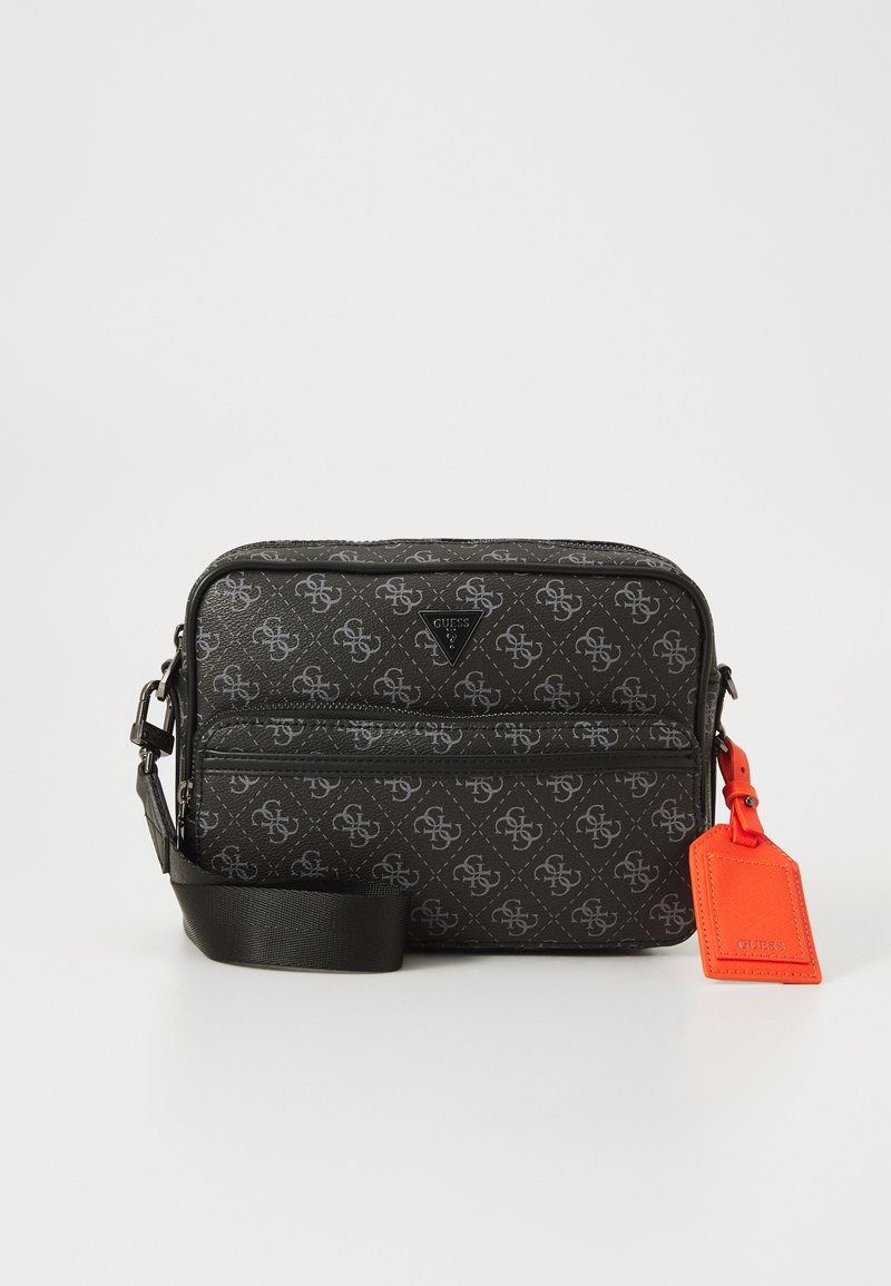 Black Guess crossbody bag with gray logo pattern, front zipper pocket, adjustable strap, and bright orange rectangular tag attached.