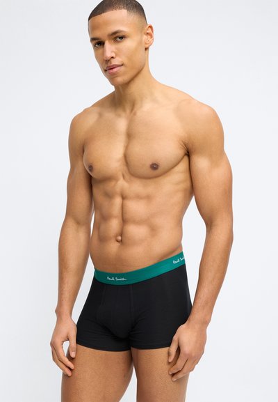 Μυώδης άνδρας φοράει μαύρα μποξέρ με πράσινη μέση που φέρει την επιγραφή "Paul Smith", στέκεται μπροστά σε ένα απλό φωτεινό φόντο.