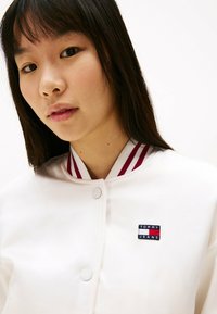 Hvid bomberjakke med maroon-stribede krave. Har et mærket Tommy Jeans-patch foran. Glat tekstur, trykknaplukning.