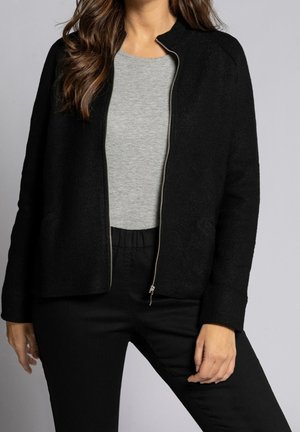 Femme portant une veste noire zippée par-dessus un t-shirt gris et un pantalon noir, debout devant un fond gris clair uni.