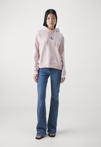 Sudadera rosa claro con un logo en el frente, capucha con cordón y puños de canalé, combinada con jeans acampanados azul y sandalias negras.