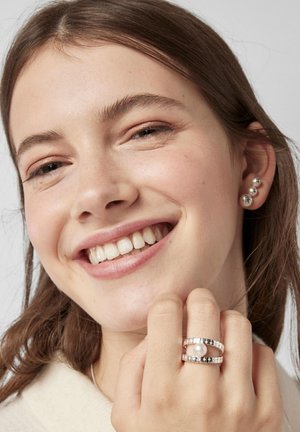 Joven sonriente, lleva un anillo de plata martillada con perla en el dedo y pendientes triples de bolas de plata, cabello castaño y liso.