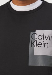 Schwarzes Baumwoll-T-Shirt mit Rundhalsausschnitt, das eine große Grafik mit dem Schriftzug "Calvin Klein" in fettem Schwarz auf weißem und grauem Hintergrund zeigt.