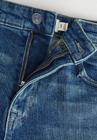 Blugi din denim cu un ton albastru, cu fermoar, hardware de bronz și o textură ușor decolorată. Au o etichetă pe talie și detalii de cusătură.