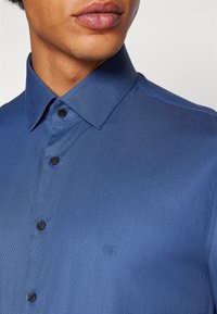 Camicia blu a trama con colletto button-down, dotata di bottoni neri e di un piccolo logo impresso sul petto.