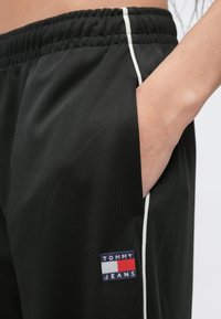 Čierne športové nohavice s elastickým pásom, bočnými vreckami a bielym lemovaním. Na nohaviciach je vyšité logo "TOMMY JEANS".