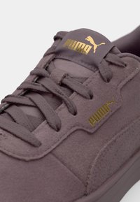 Snørresko i mørk mauve suede med et guld Puma-logo på tungen og siden, med polstret krave og teksturerede snørebånd.