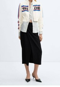 Bílá zkrácená bunda s modrými a červenými pruhy, s nápisem "PRIDE". Světlá pruhovaná košile pod ní, spárovaná s černou zavinovací sukní a špičatými botami.