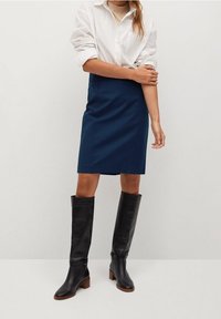 Jupe crayon bleu marine associée à une chemise blanche à boutons et des bottes noires jusqu'aux genoux avec des talons en bois. Tissu lisse, design ajusté.