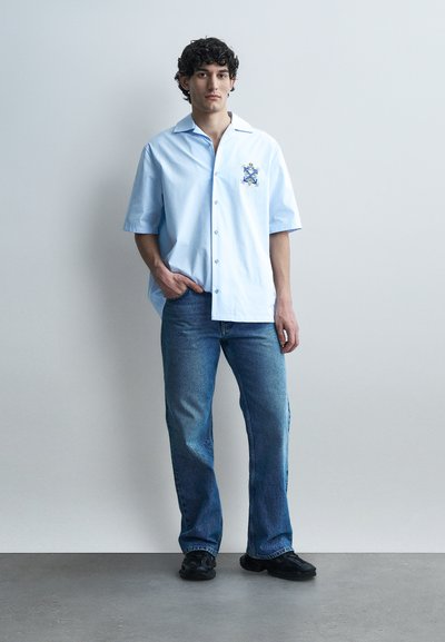 Νεαρός άνδρας όρθιος, ντυμένος με ένα ανοιχτό γαλάζιο κοντομάνικο πουκάμισο με έμβλημα και μπλε τζιν, μπροστά σε έναν απλό γκρι τοίχο.
