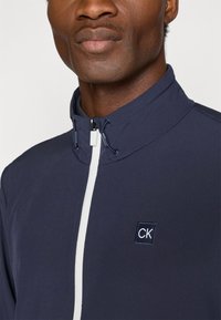 Tengerészkék kabát magas gallérral, fehér cipzárral és CK logóval. Az anyag simának tűnik, letisztult, minimalista formatervezéssel.