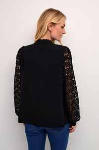 Svart blus med långa, genomskinliga spetsärmar med blommönster. Enfärgad rygg med rund nederkant. Mjuk tygtextur, avslappnad passform.