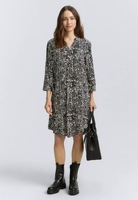 Robe noire et blanche avec un motif, manches longues, boutonnée sur le devant et coupe ample. Portée avec des bottes noires et un sac à main noir.