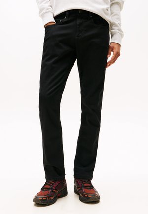 Fekete farmernadrágot és piros-fekete sportcipőt viselő személy, aki egyik kezét a zsebében tartja, másik kezét laza tartással oldalt.