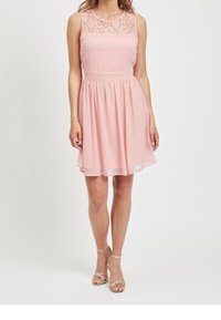 Růžové šaty bez rukávů s krajkovým zdobením, přiléhavým pasem a splývavou sukní, na postavě stojící osoby obuté v zlatých sandálech na podpatku.