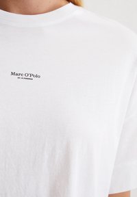 Weißes Baumwoll-T-Shirt mit Rundhalsausschnitt und kurzen Ärmeln, versehen mit einem kleinen schwarzen Logo auf der linken Brustseite. Glatte Textur.