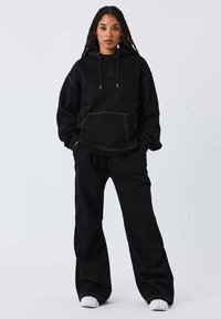 Donna con lunghi capelli intrecciati indossa un felpa nera e pantaloni larghi abbinati con sneakers bianche, in piedi contro uno sfondo chiaro e uniforme.