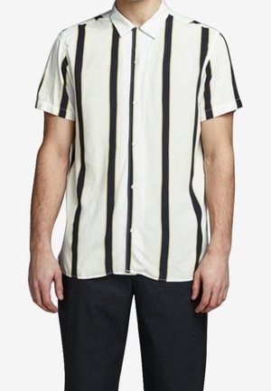 Homme portant une chemise manches courtes à boutons avec des rayures verticales noires et beiges sur fond blanc, associée à un pantalon noir uni.