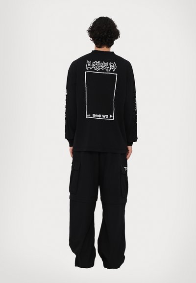 Μαύρο oversized φούτερ με γραφικά κείμενα στο πίσω μέρος και μακριά μανίκια, συνδυασμένο με μαύρο cargo παντελόνι που διαθέτει τσέπες.