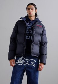 Doudoune noire avec col montant, design matelassé et détail de logo rouge. Portée sur un sweat-shirt marine et un jean en denim avec un graphique blanc.