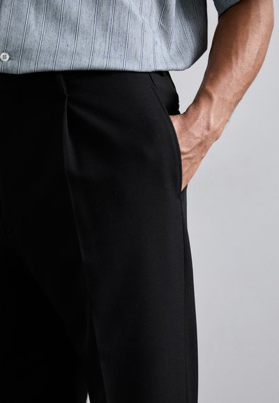 Pantaloni neri con una texture liscia e tasche laterali, abbinati a una camicia a righe azzurra chiara, parzialmente visibile. Design aderente.