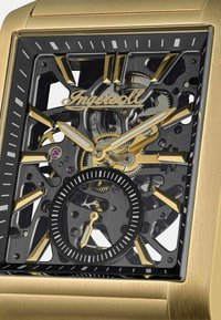 Złoty zegarek w stylu skeleton z czarną tarczą, złotymi akcentami i subtarcza. Ma prostokątny kształt i misternie wykonane detale.