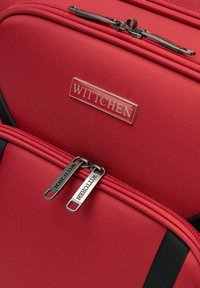 Roter Stoffrucksack mit glatter Textur, doppelten Reißverschlüssen mit Branding und einem silbernen Logo-Schild. Schwarze Akzente sorgen für Kontrast.