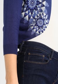 Laivastonsininen neuletakki, jossa on sinivalkoinen geometrinen kuosi olkapäällä, yhdistettynä tummiin farkkuihin, joissa on oranssit saumat.