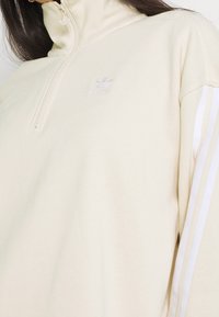 Krämfärgad sweatshirt med halvdragkedja, som har en liten broderad logotyp och vita ränder längs ärmarna.