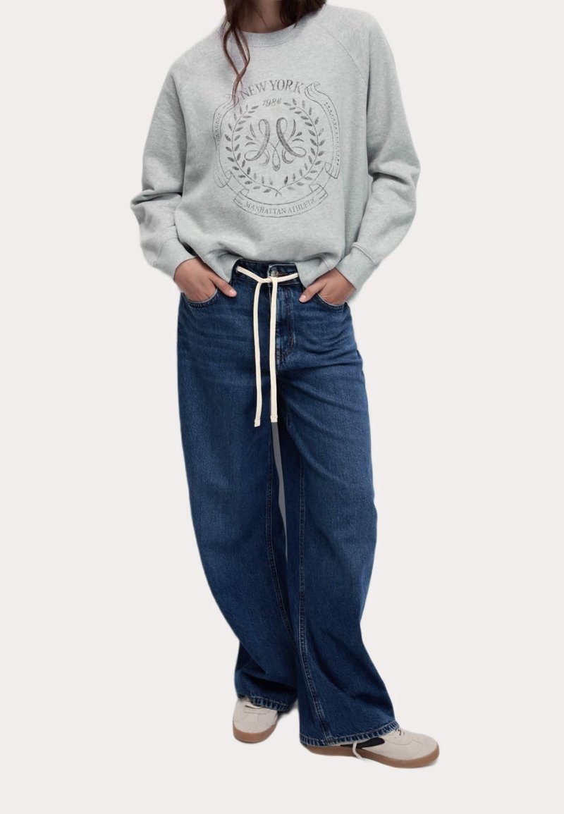Personne portant un sweat-shirt gris clair avec une inscription New York, un jean bleu ample avec un cordon blanc, et des baskets blanches, debout les mains dans les poches.
